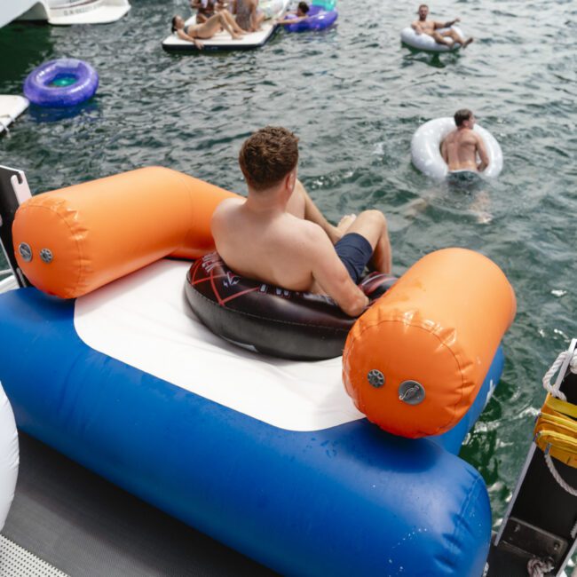 A man lounging on an orange and blue inflatable chair on a boat, with people floating on tubes in the water nearby. The scene is lively and sunny, highlighting a fun day on the water.