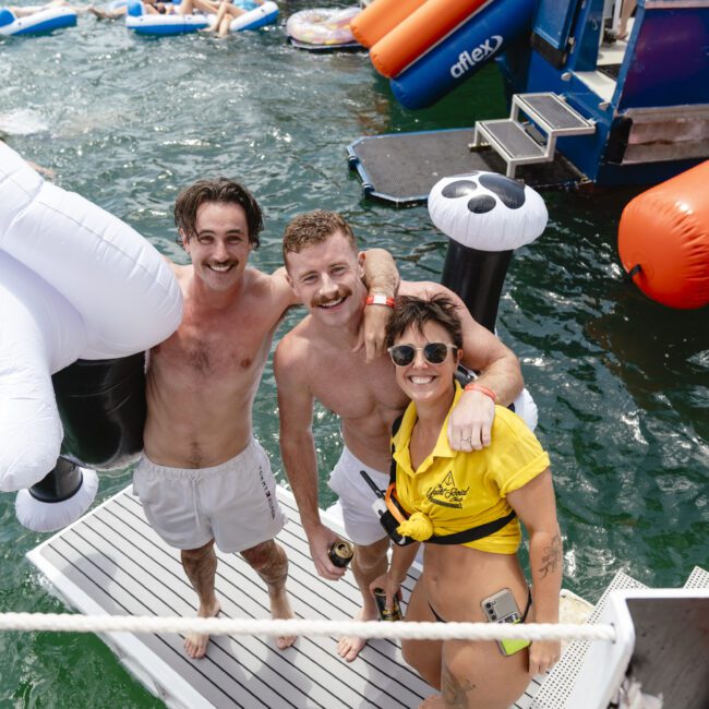 Three people enjoying a day on the water, standing on a platform by a boat. Two shirtless men smile while holding a large inflatable, and a woman in a yellow top stands beside them. Other people and inflatables are visible in the background.