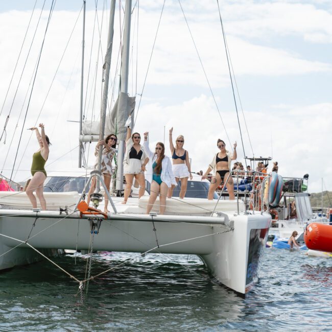 A group of people dance and pose on the deck of a sailboat on the water. Other boats are nearby, and an inflatable buoy floats in the foreground. The sky is partly cloudy.