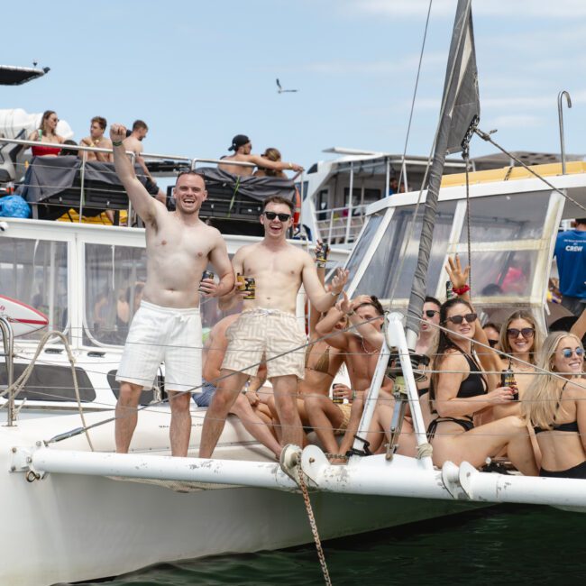 A group of people enjoy a sunny day on a catamaran, posing and smiling for the camera. Some are standing, while others sit on the boat's deck. Other boats and people are visible in the background. Everyone appears relaxed and jovial.