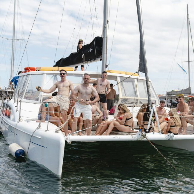 A group of people in swimsuits relax on a catamaran in the water, enjoying a sunny day. Some individuals are sitting, while others stand on the boat's deck. Other boats are visible in the background.