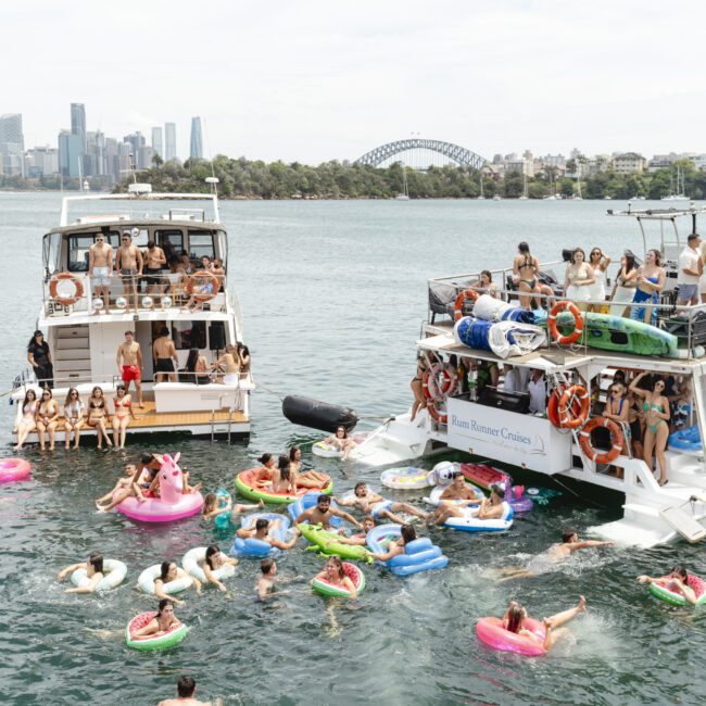 A lively scene on the water with two boats filled with people enjoying a sunny day. Many are swimming and floating on inflatable toys, with a city skyline and bridge visible in the background.