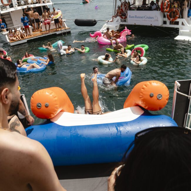 People enjoying a party on a lake with inflatable floats. A person is doing a handstand on an inflatable slide, while others relax on floaties nearby. Boats are anchored in the background with more people on board.