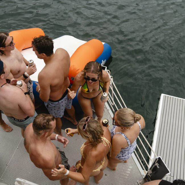 A group of people in swimwear are gathered on a boat's deck, holding drinks and chatting. The deck is next to a floating mat on the water. The atmosphere is relaxed and festive.