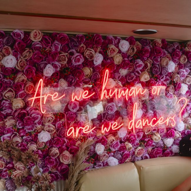 A wall covered in pink and purple roses features a neon sign that reads, "Are we human or are we dancers?" Below is a beige sofa with a person's arm resting on it. Dried plants are placed next to the sofa.