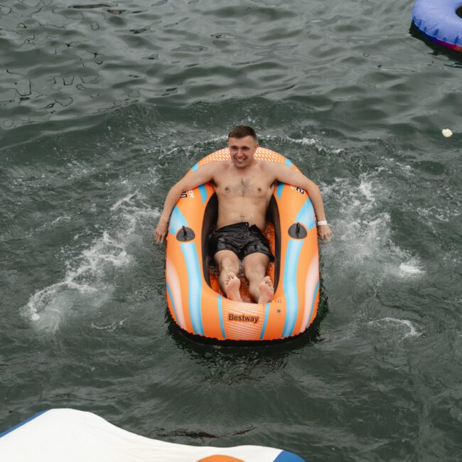 A shirtless person is relaxing in an orange and white inflatable ring on a body of water. Other inflatable rings float nearby. The water surrounding them is dark, and they appear content.