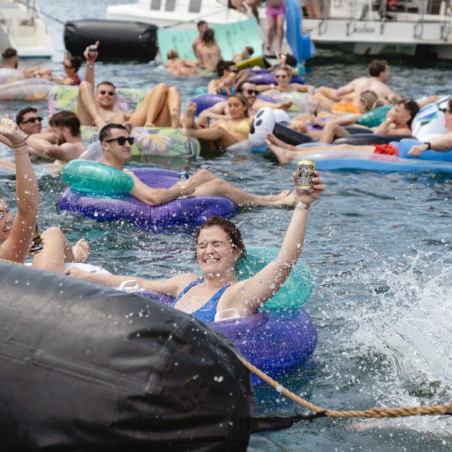 A group of people enjoying a party on a lake, sitting on inflatable rafts and inner tubes. They're surrounded by water, with boats in the background. One person is raising a can in celebration, and others are relaxing under the sun.