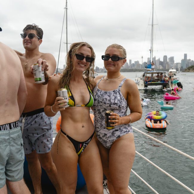 A group of people in swimwear stands on a boat, holding drinks and smiling. In the background, other boats and inflatables are visible on the water, with a city skyline under a cloudy sky.