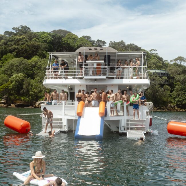 People enjoy a sunny day on a boat with multiple decks and water slides. Some are lounging on inflatables in the water, while others are on the boat. The scene is set against a backdrop of lush greenery.