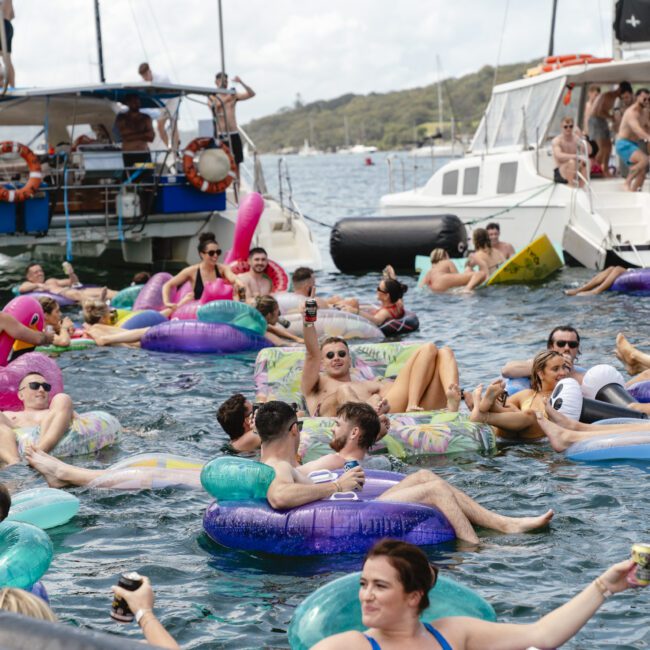 A group of people relax on colorful inflatable floats in the water, surrounded by boats. They are enjoying a sunny day, holding drinks, and having a fun time together.