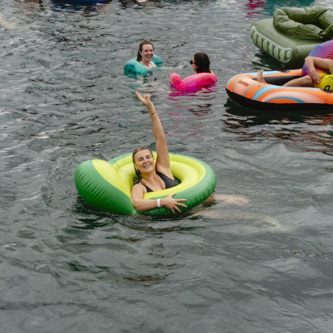 A woman in a green float joyfully raises her arm in a body of water, surrounded by other people in colorful inflatables. The atmosphere is lively and playful.