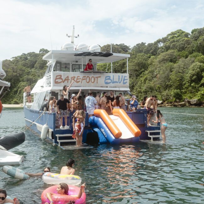 A group of people are enjoying a day on a large boat named "Barefoot Blue" anchored near a lush, green shoreline. Some are on an inflatable slide attached to the boat, while others relax on colorful floaties in the water.