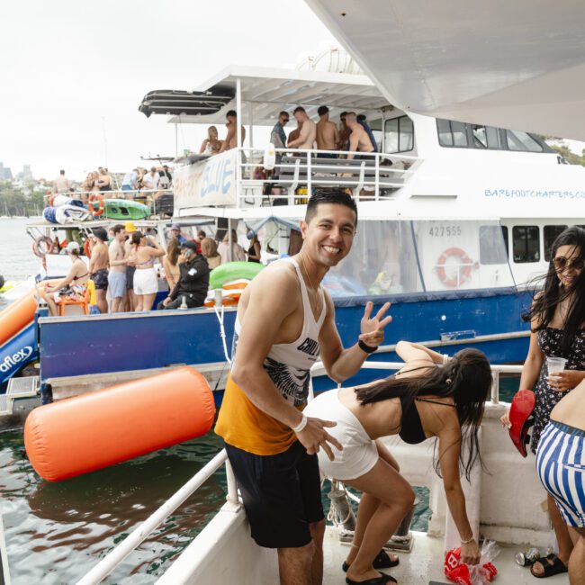 A man in a tank top smiles and gives a peace sign on a boat. In the background, people are gathered on another boat having a party. The scene is lively, with orange flotation devices visible near the boats.