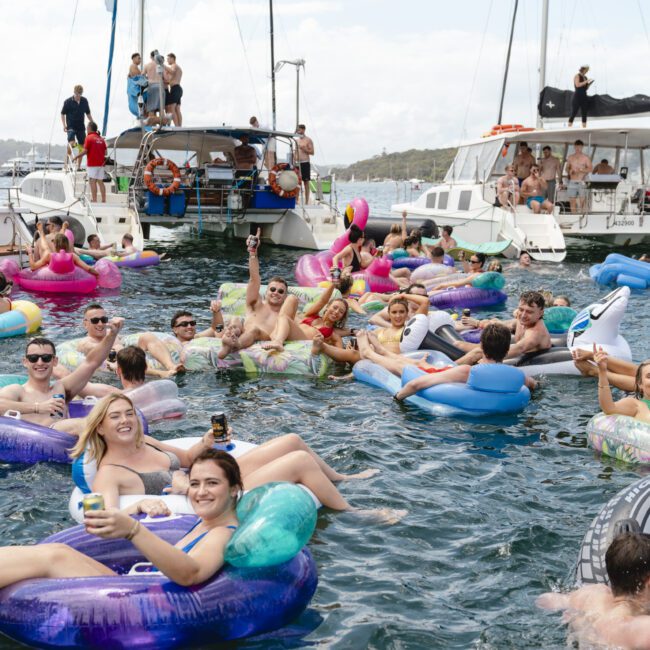 People are enjoying a sunny day on the water, floating on colorful inflatable tubes. Several boats are anchored nearby, and some individuals are on the decks. The scene is lively and festive, with everyone appearing relaxed and cheerful.