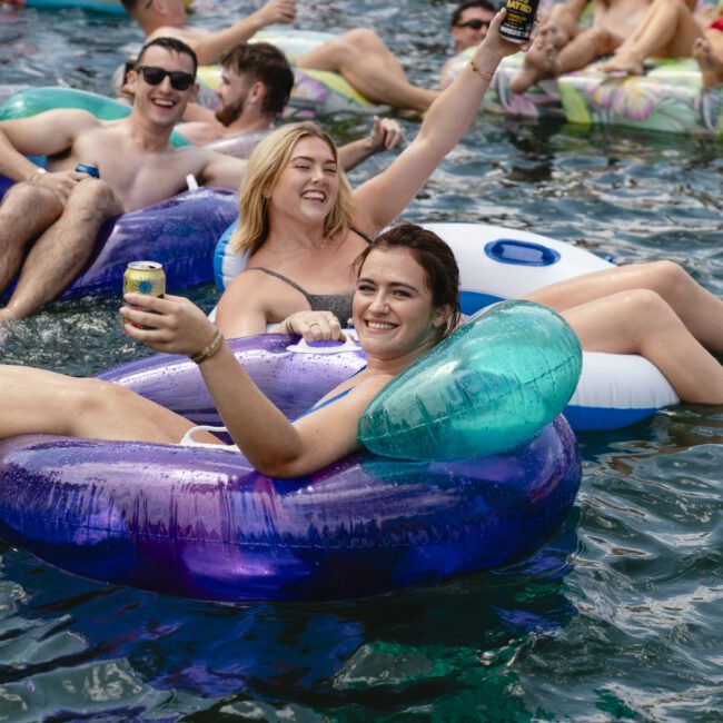 A group of people enjoying a sunny day, floating on inflatable tubes in a body of water. They are smiling, raising drinks, and relaxing in swimwear. The scene is lively and cheerful.