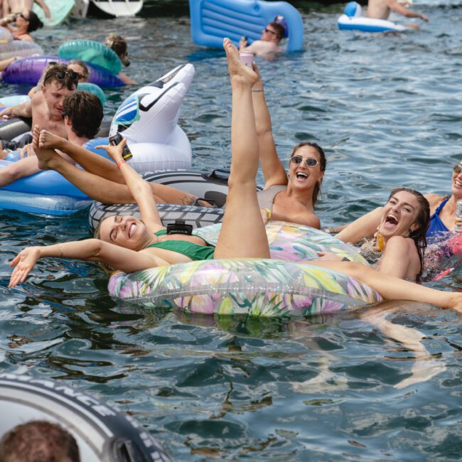 A group of people in swimsuits enjoy floating on inflatable rafts and toys in a lake. They are smiling and laughing, holding drinks, and relaxing in the water on a sunny day.