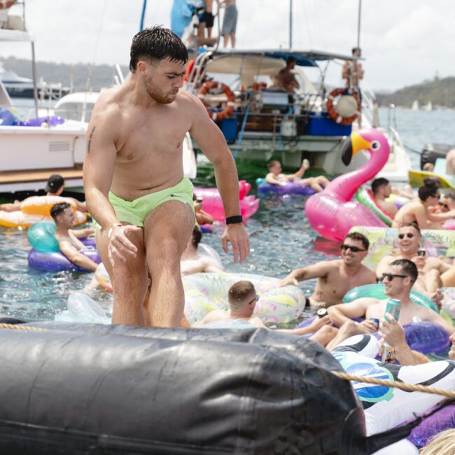 A group of people enjoying a lively party on the water with colorful inflatable toys, including a large pink flamingo. Boats are anchored nearby, and the atmosphere is festive and vibrant under a clear sky.