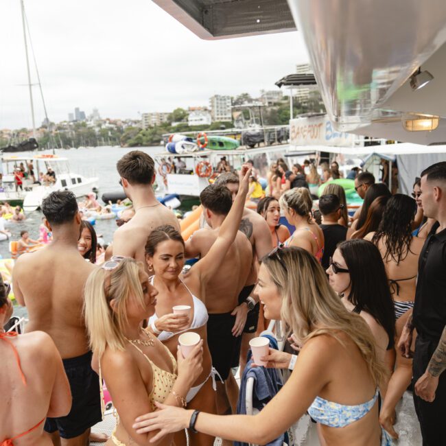 A lively scene on a boat with a crowd of people in swimsuits enjoying a party. Some are holding drinks, and others are socializing. Boats and inflatables are visible in the water nearby. The atmosphere is festive and relaxed.