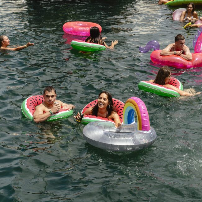 People enjoying a sunny day in the water with colorful inflatable rings shaped like watermelon slices and unicorns. The scene is lively and playful, with individuals scattered across the water, surrounded by green, pink, and silver floats.