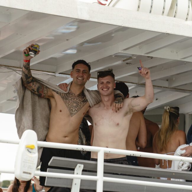 A group of people on a boat party, celebrating and having fun. Two shirtless men in the foreground smile and pose with drinks, while others mingle in the background. The atmosphere is lively and festive.