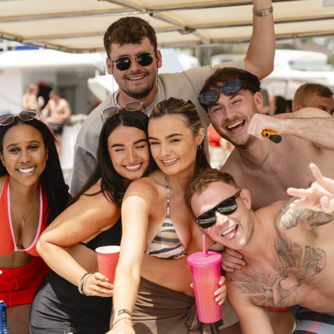 A group of six people on a boat, smiling and posing for the camera. They are casually dressed for warm weather, with some holding drinks. The background shows a sunny day with a beach setting.