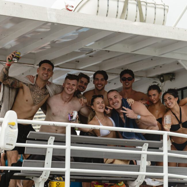 A group of people in swimwear enjoying a boat party. They are smiling, holding drinks, and posing together under a shaded area on the boat.