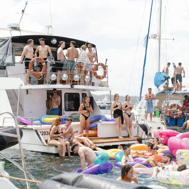 A lively crowd enjoys a party on two docked boats. People are mingling on the decks and floating on colorful inflatables, including a pink flamingo, in the water. It’s a sunny day and the atmosphere is festive and relaxed.