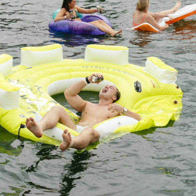 A man relaxes on a yellow inflatable raft shaped like a pineapple, drinking from a can while floating in a body of water. Two other people on inflatable loungers are nearby, enjoying the water as well.