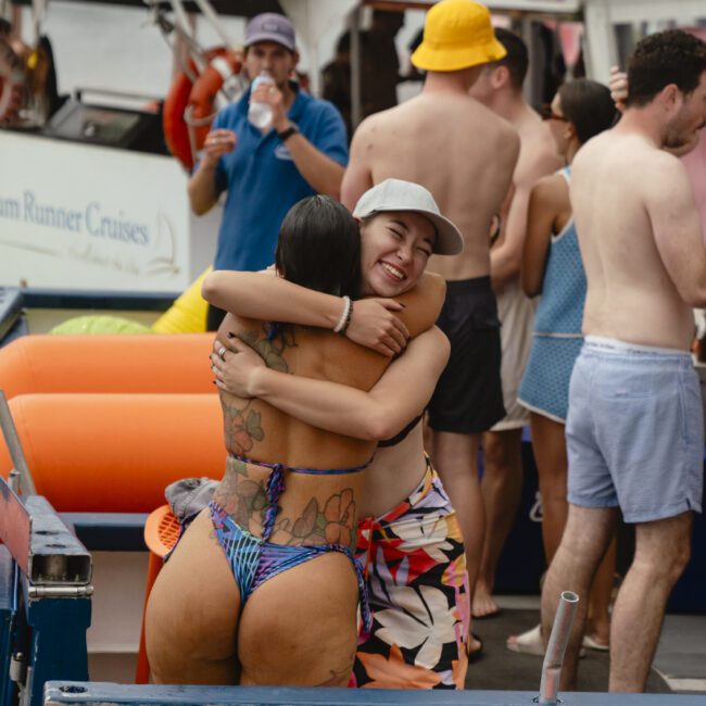 Two people hugging on a boat deck, surrounded by others enjoying a casual gathering. One person is wearing a swimsuit, and the other has a sun hat and colorful wrap. The background features more people, life vests, and boating equipment.