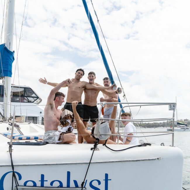 Five people are happily posing on a sailboat named Catalyst. They are wearing swimwear and smiling, with arms raised. The background shows calm waters and a coastal skyline under a partly cloudy sky.