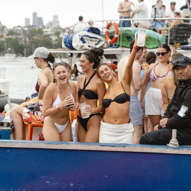 A group of people enjoying a pool party on a boat. Three women in swimwear are smiling and holding drinks, while others around them relax and socialize. The background shows additional boats and a city skyline in the distance.