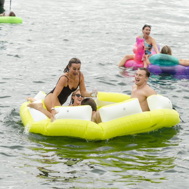 A group of people enjoying a sunny day on a lake. Several individuals are lounging on bright inflatable floats, including a yellow one in the foreground. Others are on various colorful floats in the background, smiling and interacting.