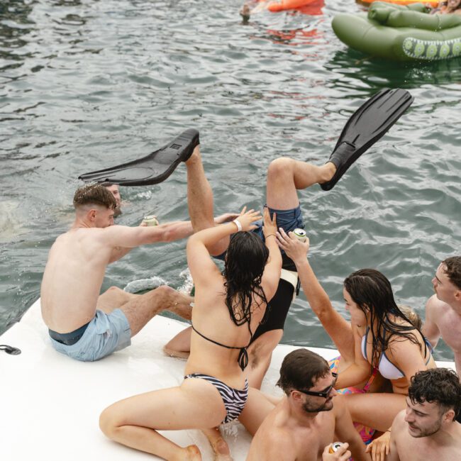 People are enjoying a day on the water with inflatable floats. Some are wearing swimsuits and holding drinks, while others are playfully interacting. A woman on a floating platform appears to be helping someone who's in large flippers.