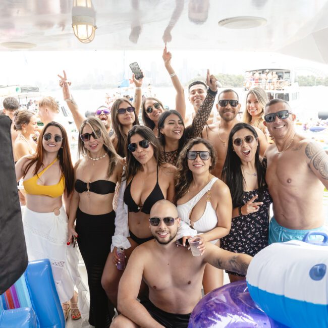 A group of people in swimwear are smiling and posing together on a boat during a sunny day. They are wearing sunglasses and appear to be enjoying a party atmosphere, with inflatable items visible in the background on the water.