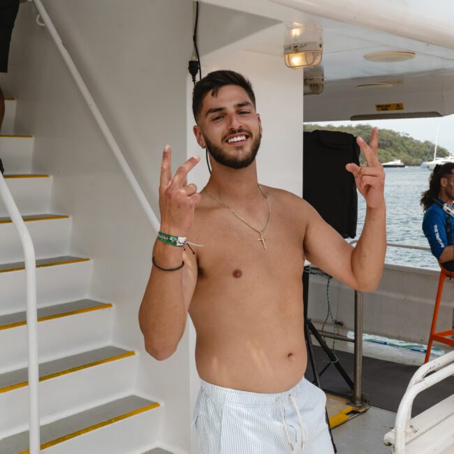 A man in swim trunks stands on a boat, smiling and making peace signs with both hands. The boat's interior and stairs are visible, with the ocean and hills in the background. It's a sunny day.