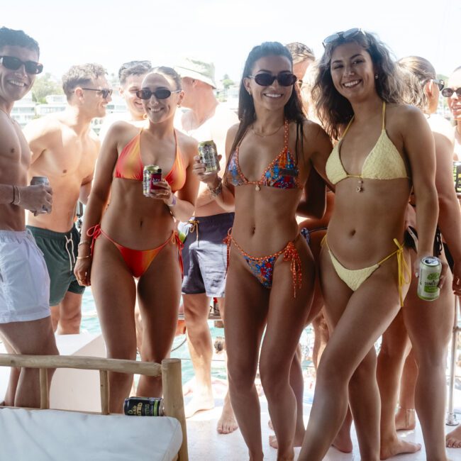 A group of people enjoying a sunny day on a boat. Some are holding drinks, and several are wearing swimwear and sunglasses. The background shows water and blurred greenery, indicating they're near a shore or harbor.