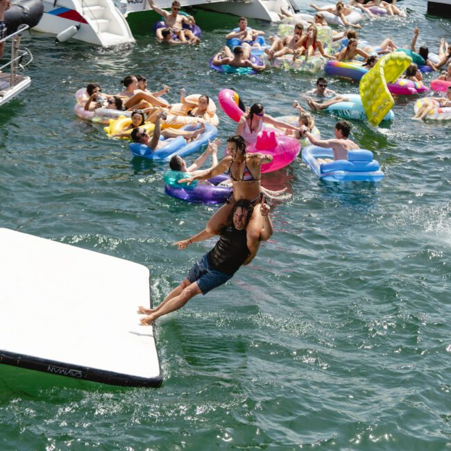 People are enjoying a sunny day at a crowded lake, floating on colorful inflatables. A man jumps into the water from a floating platform, carrying a woman on his shoulders. Several boats are docked nearby.