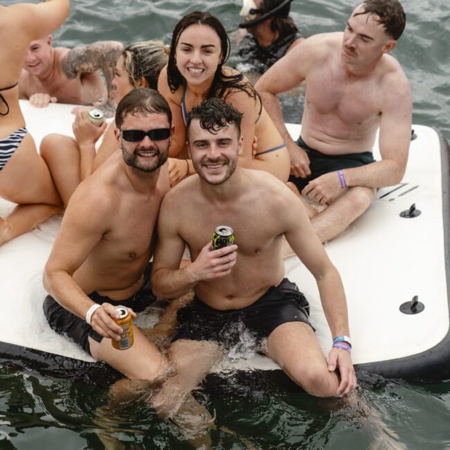 A group of people enjoying a day on the water, sitting and standing on a floating platform. Some are holding drinks, and all are in swimsuits, smiling and relaxed in the sunny setting.