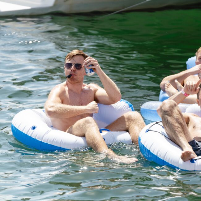 Three men are lounging in inflatable tubes on a body of water, enjoying a sunny day. One man is drinking from a can, while another raises his arm with a playful expression. All appear relaxed and content.