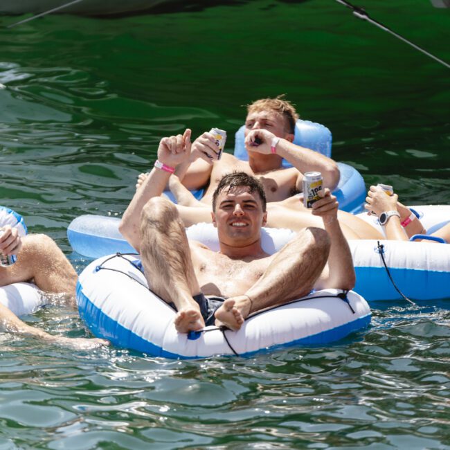 A group of young adults relaxing and drinking on inflatable tubes in a lake, all wearing swimsuits and enjoying a sunny day. The water is calm, and they're surrounded by green, summery scenery.