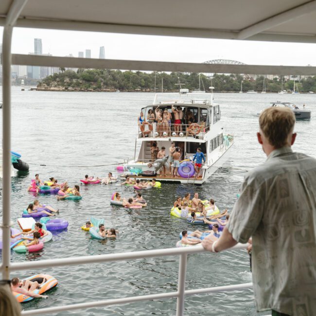 A person stands on a boat's deck overlooking a lively scene of people swimming and floating in the water on colorful inflatables. Several boats are anchored nearby. The city skyline is visible in the background under a cloudy sky.