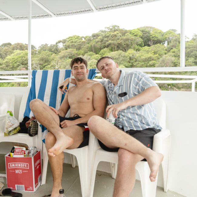 Two men sitting on a boat deck, one shirtless and the other in a striped shirt, relaxing. A box of beer and food wrappers are on the floor. Dense greenery is visible in the background.