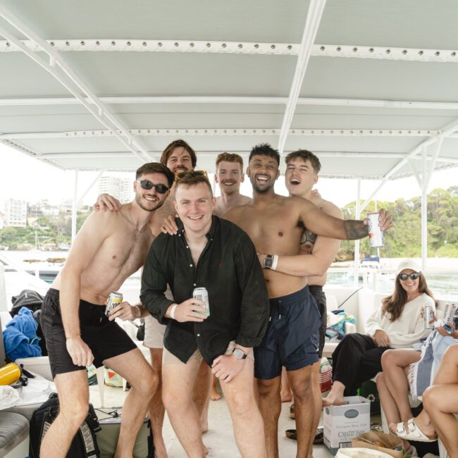 A group of friends poses for a photo on a boat, smiling and holding drinks. Some are wearing swimwear and casual clothing. They are surrounded by interior seating and natural scenery is visible in the background. Happy and relaxed atmosphere.