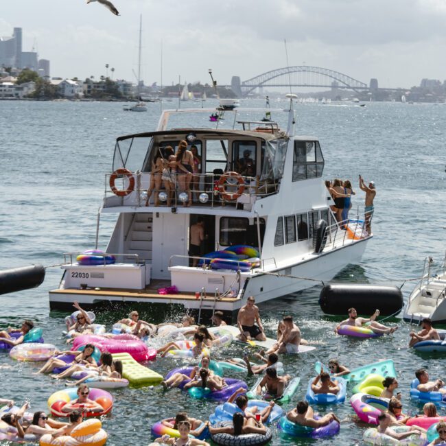 A lively scene on a sunny day features a boat on a bustling harbor, surrounded by numerous people relaxing on colorful inflatable floats in the water. The skyline and a prominent bridge are visible in the background.