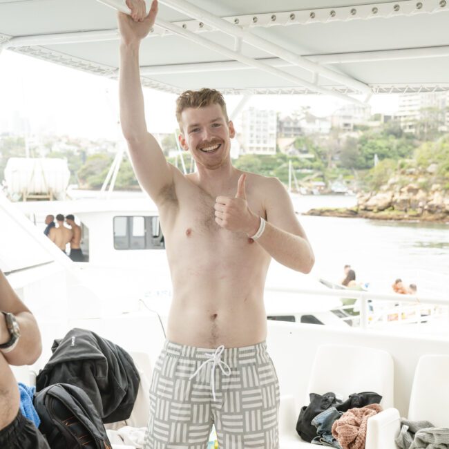 A man in patterned swim trunks gives a thumbs up while standing on a boat. He is smiling and under a canopy, with boats and a rocky shoreline visible in the background.