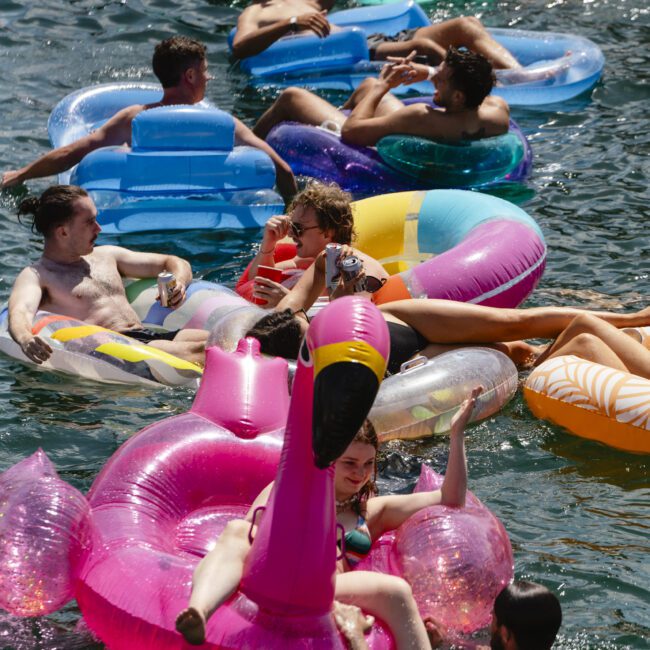 A group of people relax on colorful inflatable floats, including a pink flamingo, in a body of water on a sunny day. The scene is lively, with individuals socializing and enjoying the sunshine.