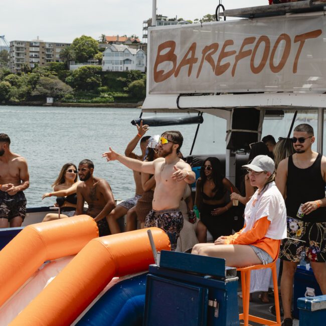 A group of people enjoys a day on a boat with a slide, wearing swimwear. Some are dancing, others are sitting or taking pictures. The background shows houses, trees, and water under a clear sky.