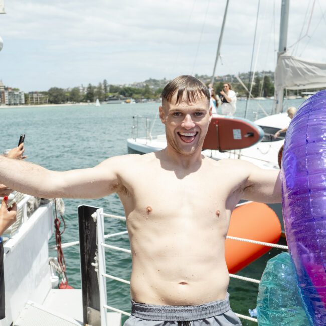 A smiling shirtless man stands on a boat deck holding an inflatable purple ring. He is surrounded by people and the ocean is visible in the background with sailboats and distant buildings. The day is sunny and bright.