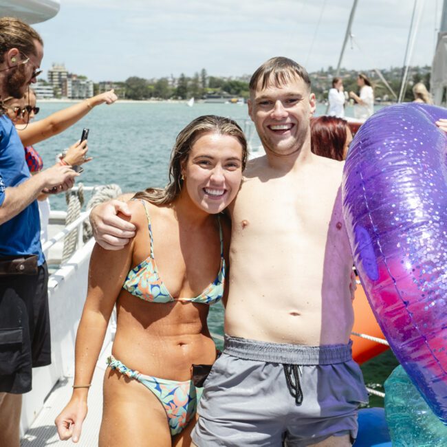 A smiling woman in a bikini and a shirtless man with a large purple float stand together on a boat. Other people are visible in the background near the water. The scene is sunny, with buildings visible in the distance.