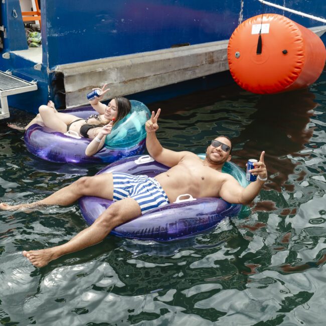 Two people relax on inflatable tubes in the water near a boat. One, wearing a bikini, holds a can and smiles, while resting their head. The other, in striped trunks, holds a drink and gestures peace signs. A bright orange buoy floats nearby.